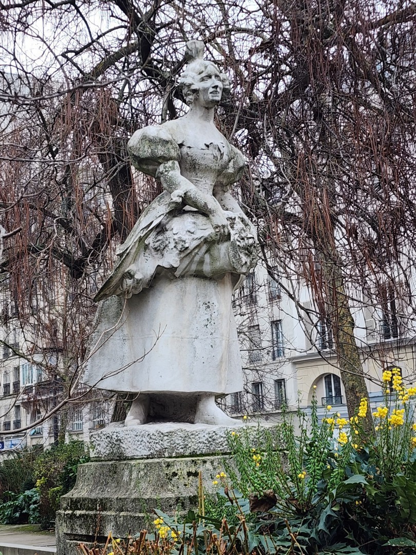 Statue d'une jeune femme du 19eme siècle vêtue d'un grossier habit gris dans lequel elle a mis des fleurs qu'elle propose aux passants
