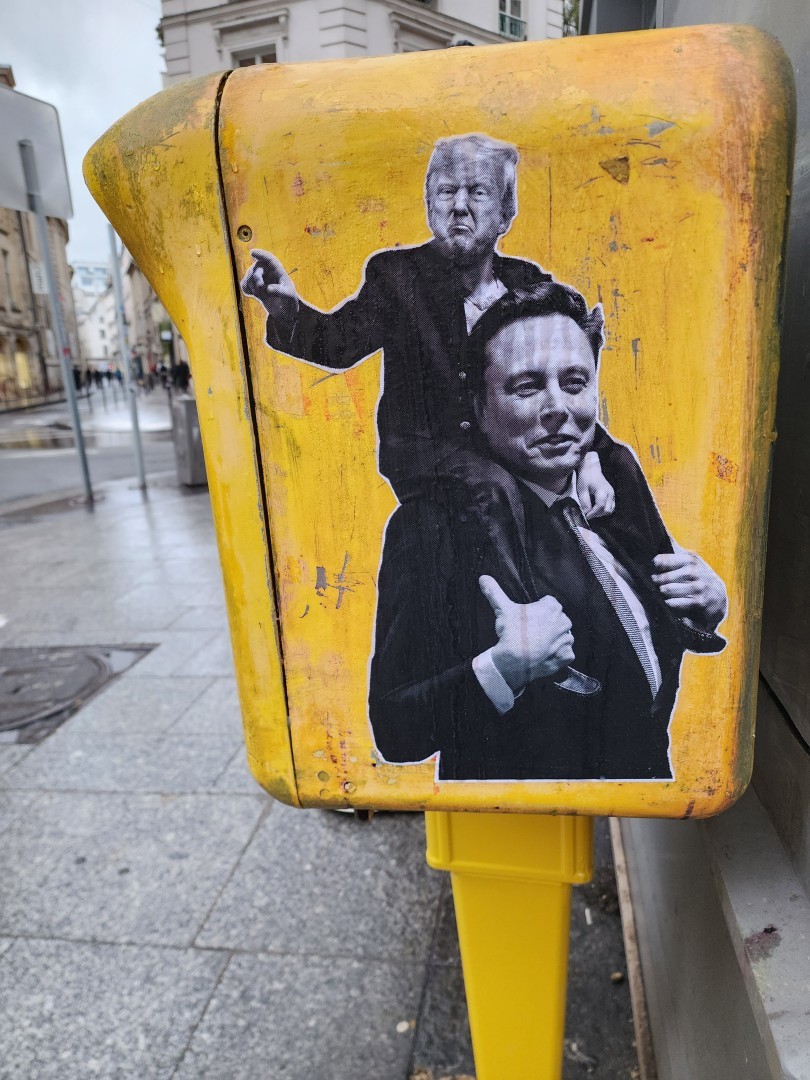 On a Paris' mailbox, Trump is ridding a hugely smiling dumb Musk. Collage in black and white
