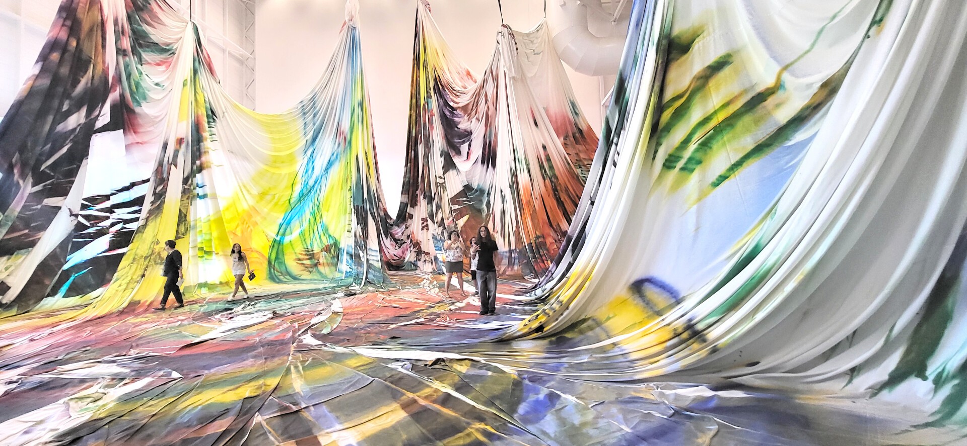Draperies colorées accrochées au plafond de la Grande Nef du Centre Pompidou Metz. Au centre est ménagée une clairière plane, au bord de laquelle deux personnes se font face.
