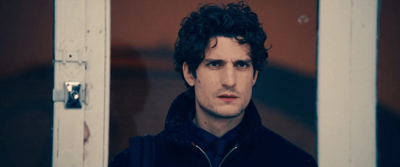 L'acteur Louis Garrel, habillé de noir, au centre de l'image, l'air concentré. Derrière lui, une fenêtre laisse passer une lumière orange sombre