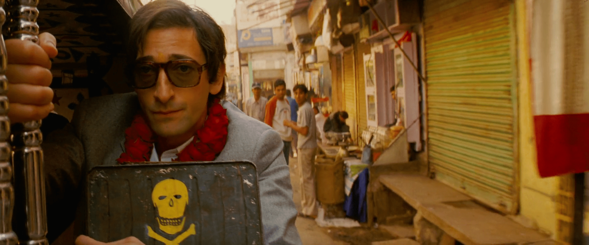 Adrien Brody, collier de fleurs rouges autour du cou et lunettes de soleil, se tient en partie hors d'un moto-taxi dans une rue en Inde, il tient une boite sur laquelle il y a une tête de mort.