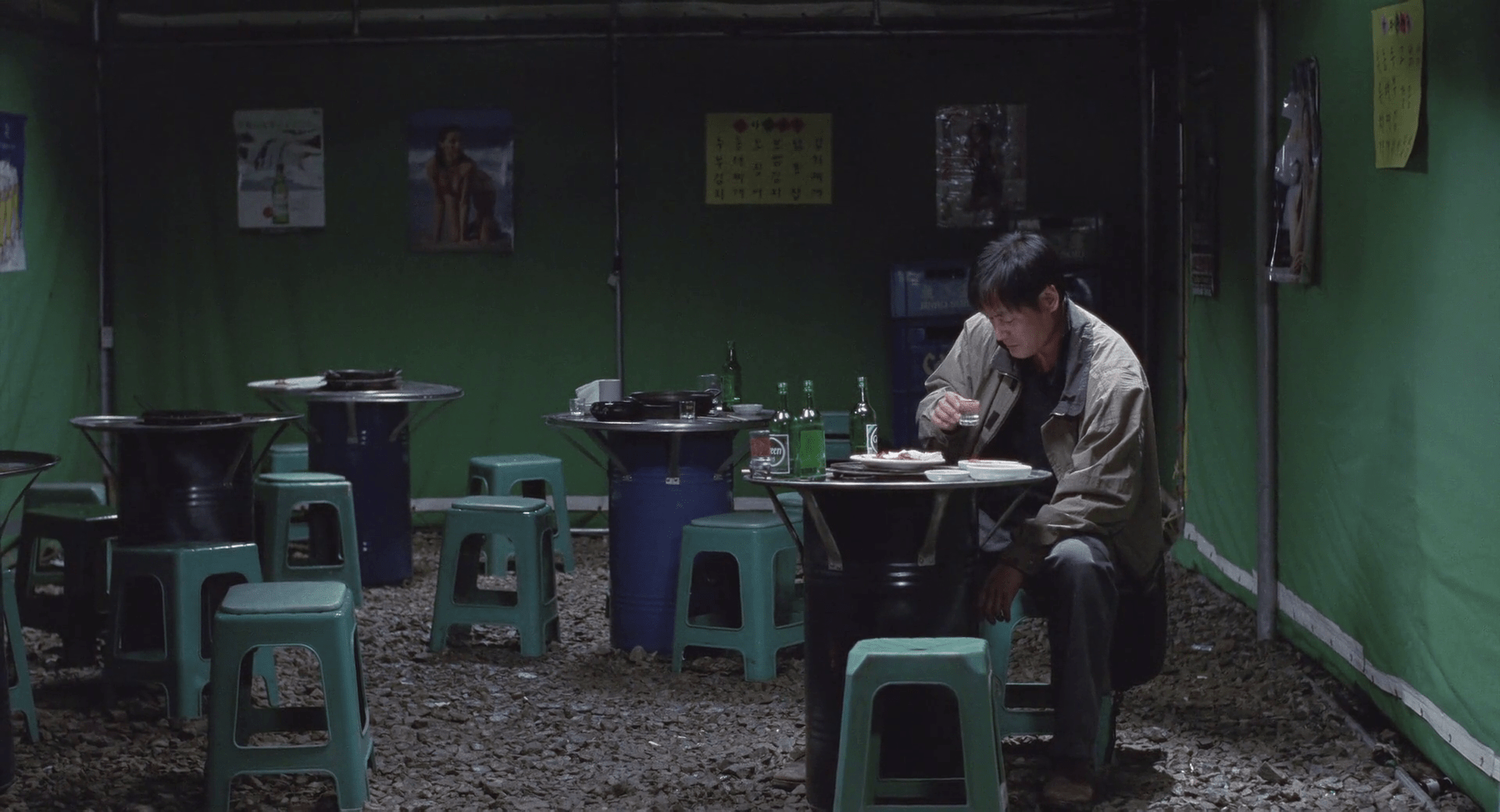 Une salle de fête sous chapiteau vert, avec quelques petites tables rondes, une personne assise, sur le point de porter un verre à sa bouche, le regard triste et le corps courbé, portant une veste verte