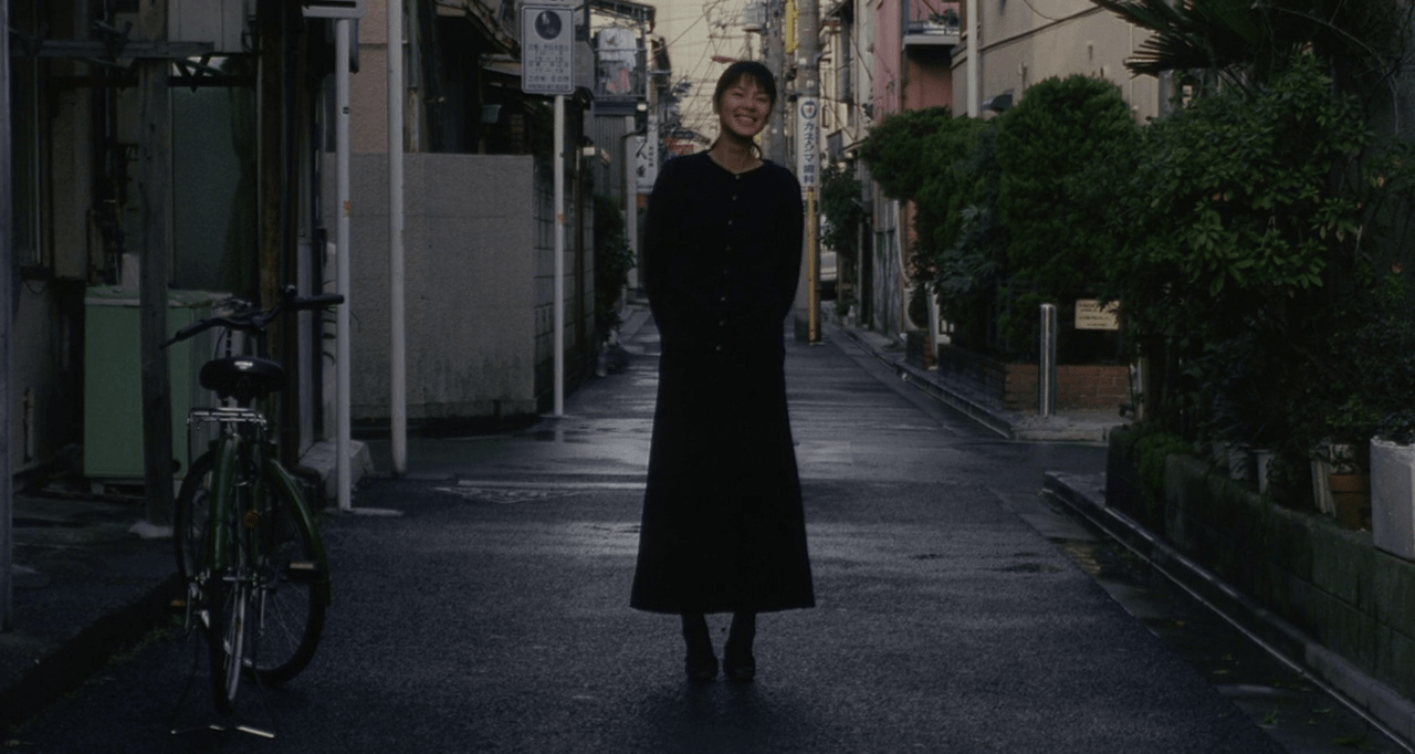 L'actrice Makiko Esumi, toute de noire vêtue, se tient debout, les mains dans le dos, le visage souriant, au milieu d'une rue détrempée, un vélo vert sur le côté.