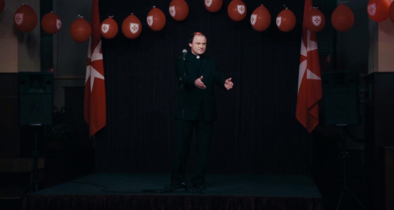 Une petite scène sombre, entourée de 2 baffles. Des ballons gonflables et 2 drapeaux rouges avec une croix blanche décorent la scène. Un prètre debout sur la scène, derrière un micro, tend les bras comme pour accueillir quelqu'un.