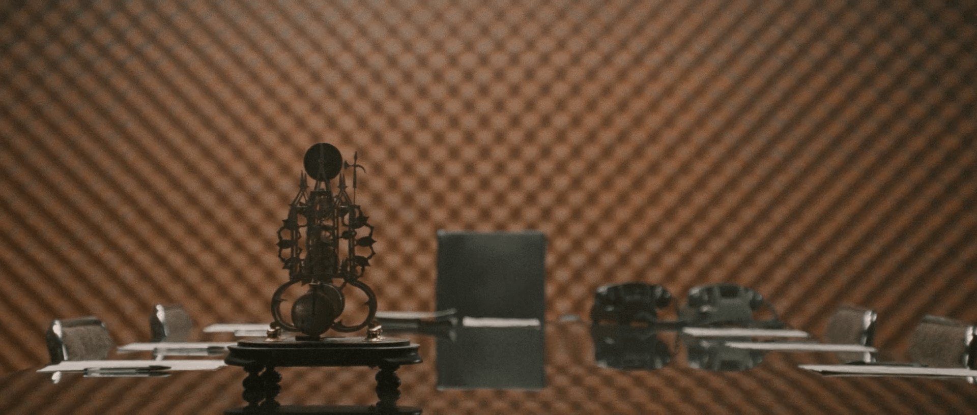 Un bureau en bois, brillant. Une horloge antique posée dessus, presque au centre. Quelques documents, deux téléphones posés sur la table. Les murs sont orange-bruns, avec un motif rectangulaire (c'est un mur insonorisé)