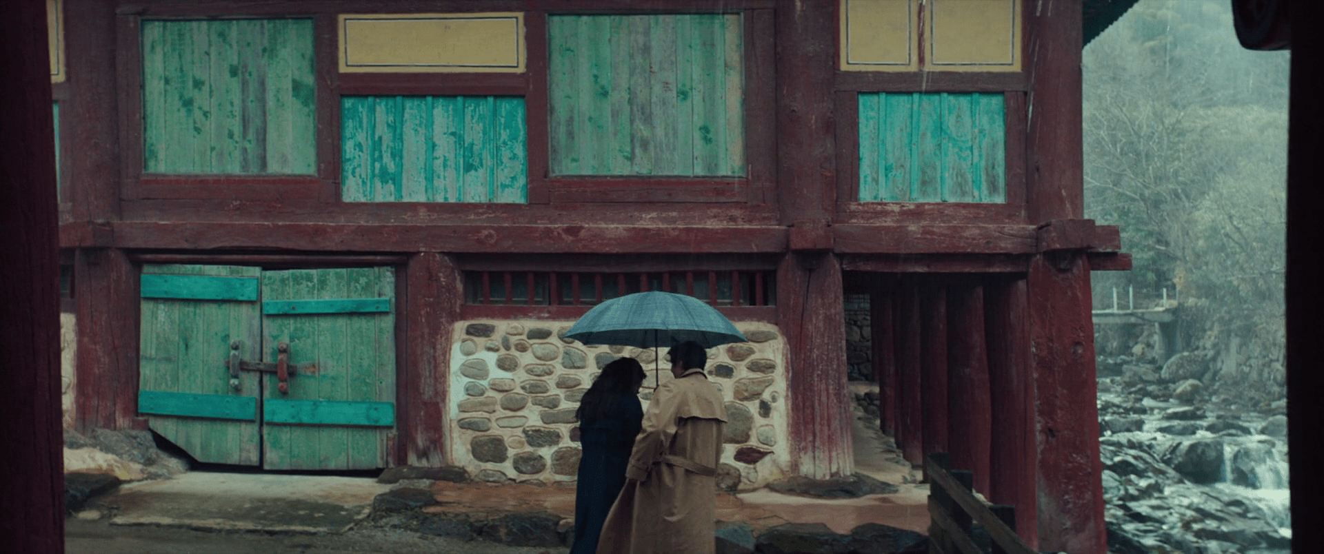 Une grande maison en bois rouge, avec des portes et volets verts et jaunes. A l'arrière-plan sur la droite, une rivière pleine de rochers. A l'avant-plan, de dos, une femme en bleu et un homme de manteau beige lui tenant un parapluie.