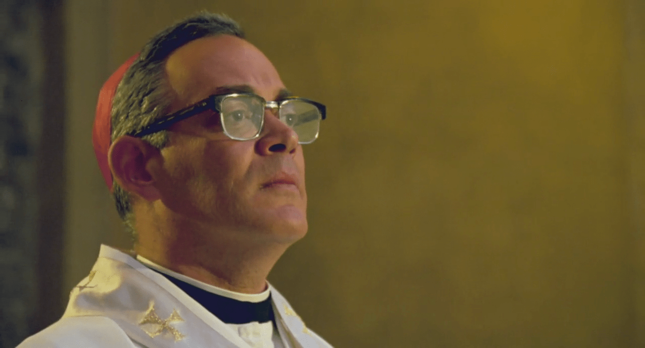 Un prêtre (Raul Julia), portant des lunettes, regarde fixement sur la droite. Derrière lui, un mur jaune.