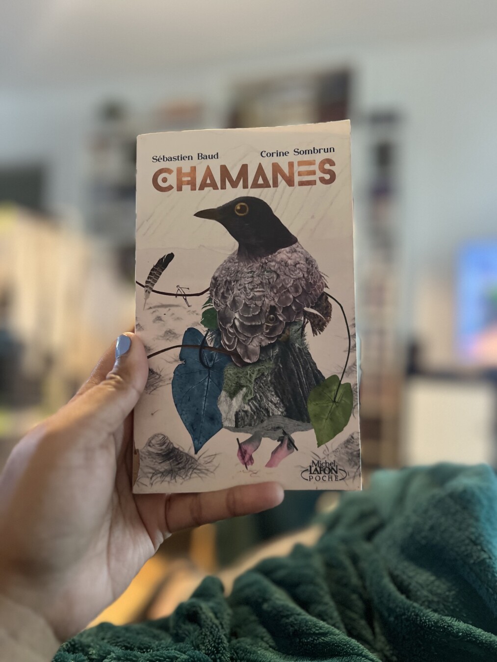 Je tiens dans ma main un livre. On voit la Couverture du livre Chamanes de Sebastien Baud et Corine Sombrun. La couverture représente une corneille esthétisée avec des feuilles et des mains.