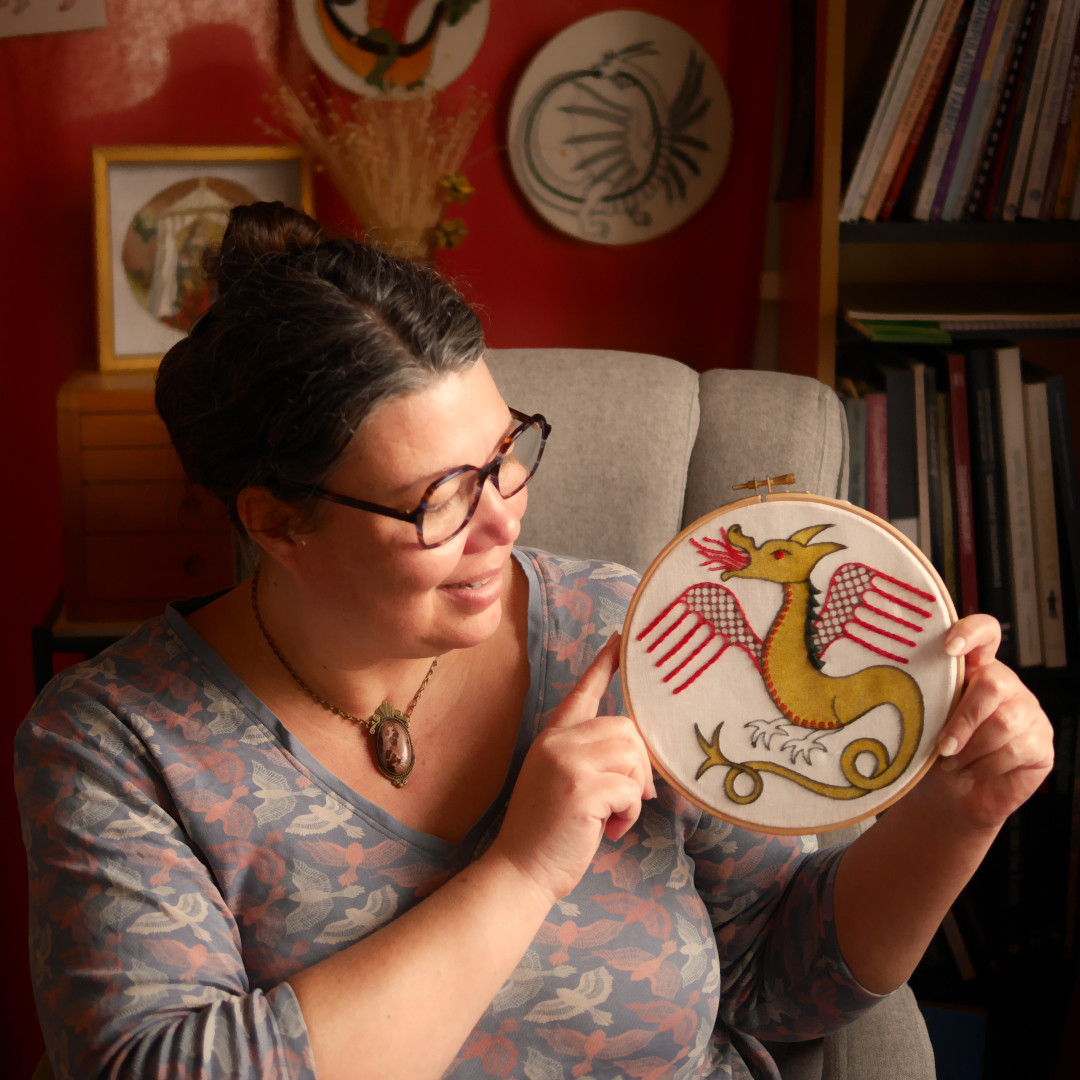 Portrait de moi, femme brune qui tient dans ses mains un tambour de broderie avec un dragon médiéval brodé en rouge et vert sur fond imprimé doré