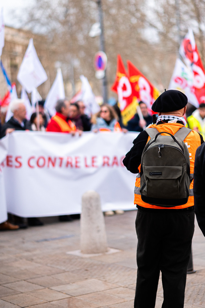 Une personne avec un gilet orange (CFDT), un kéfié et un béret, prend en photo la tête de cortége