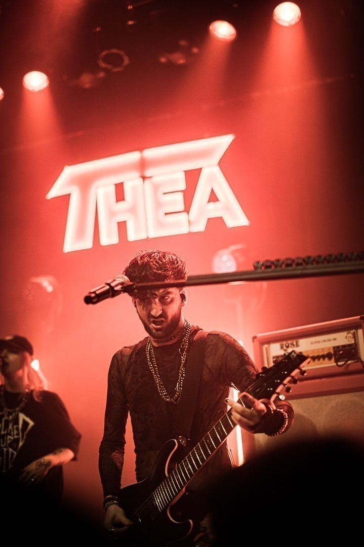 Le guitariste qui à l'air énervé, le tout sous une lumière rouge avec THEA écrit en gros au dessus