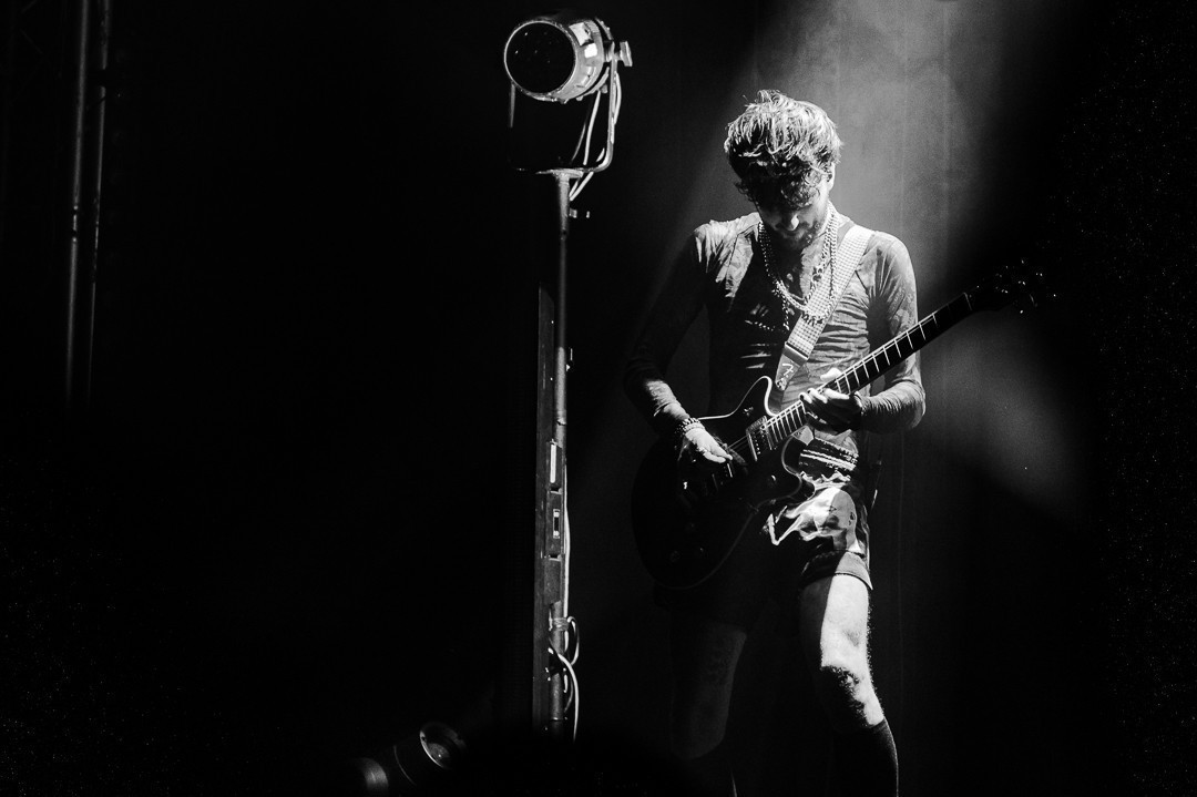 Noir et blanc, le guitariste joue la tete baissé, éclairé par des spots, faisant apparaitre des ombres marqué sur lui