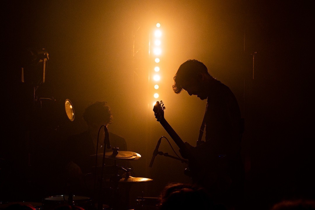 Photo sombre baigné dans une ambiance jaune orangé, on voit la silhouette du guitariste et on perçois le batteur