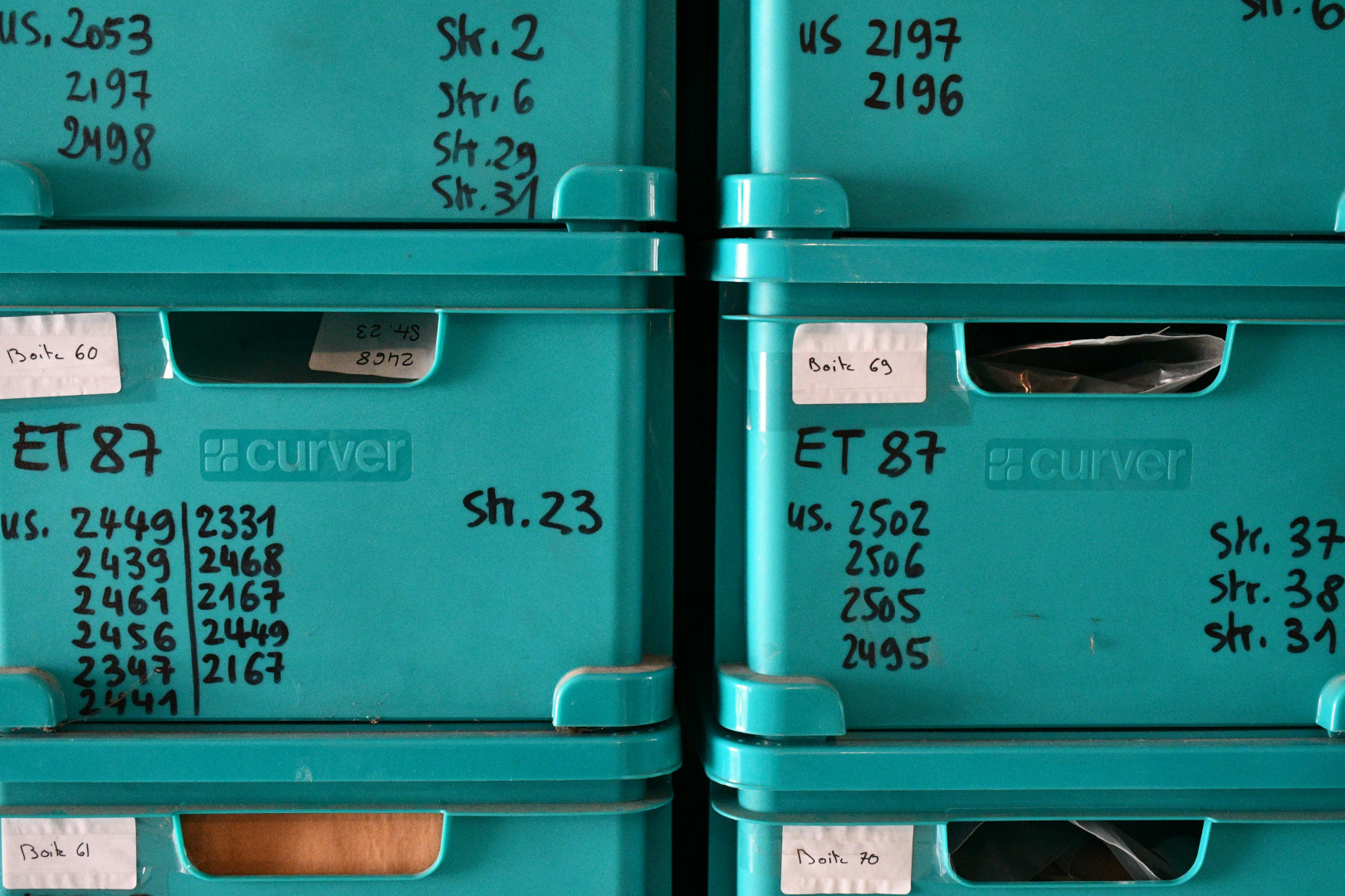 La photo montre des six caisses de plastique vert turquoise empilées les unes sur les autres, portant la mention de ce qu'elles contiennent inscrit au feutre noir.