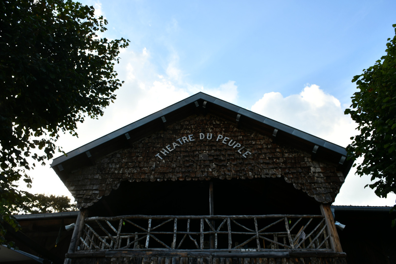 Sur un fond de ciel bleu traversé par un gros nuage blanc, se détache la silhouette du pignon d'un bâtiment de bois, semblable à un chalet. Sur le haut du pignon est inscrit Théâtre du Peuple en lettre capitales blanches. La photo est encadrée d'un arbre sur chacun de ses côtés.