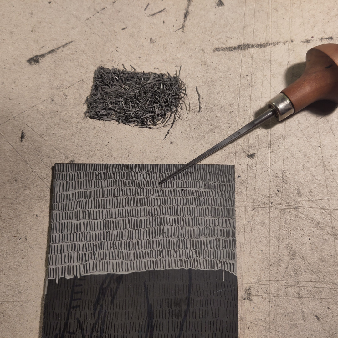 photo vue de dessus d'une petite plaque de caoutchouc gravée à un peu moins de la moitié 
une gouge très fine est posée à côté, la pointe sur la plaque
au dessus de la plaque sur le bureau un petit tas bien rangé des chutes de caoutchouc enlevée lors de la gravure