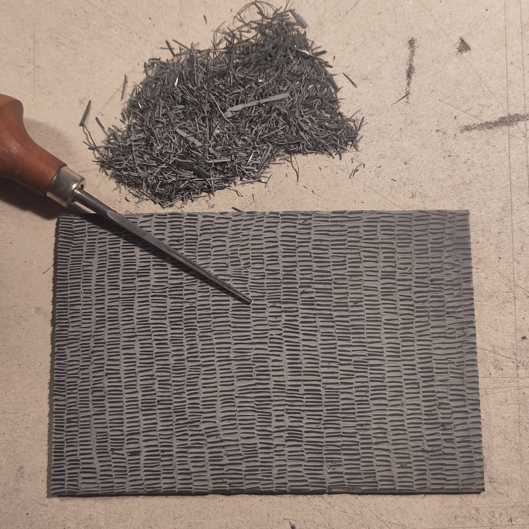 photo prise de dessus d'une petite plaque de caoutchouc entièrement gravée de petits traits verticaux.
une gouge fine est à moitié posée dessus. un petit tas de copeaux de caoutchouc est rassemblé à côté