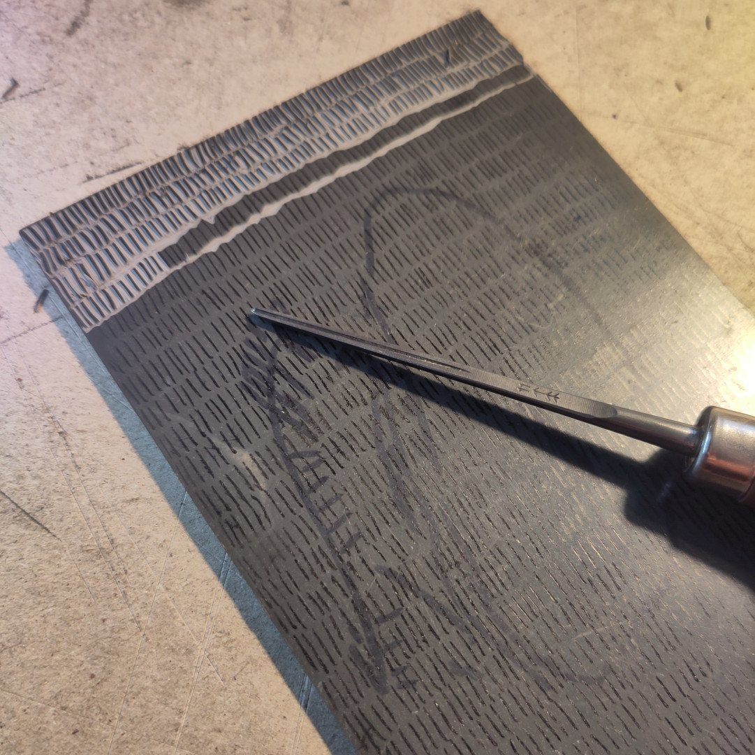 photo en gros plan d'une plaque de caoutchouc en cours de gravure.
le motif c'est des rangées de petits traits verticaux tracés au stylo plume, sur toute la surface de la plaque
une gouge très fine est posée dessus