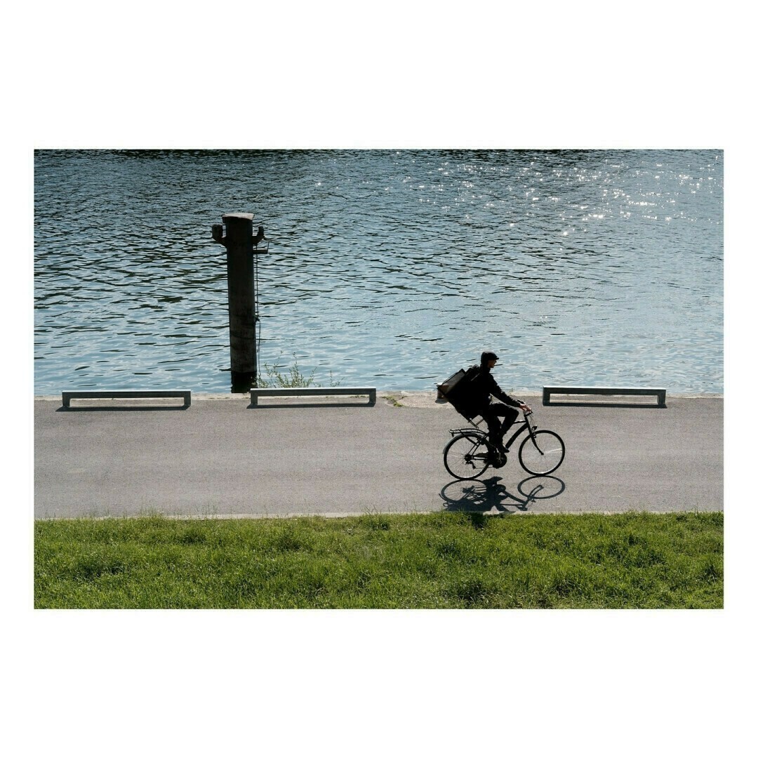 Un homme en tenue sombre, avec un grand sac de livraison sur le dos, roule sur une piste au bord de l’eau. Son ombre s’étire sur le sol tandis que la lumière du soleil scintille sur la surface de l’eau.