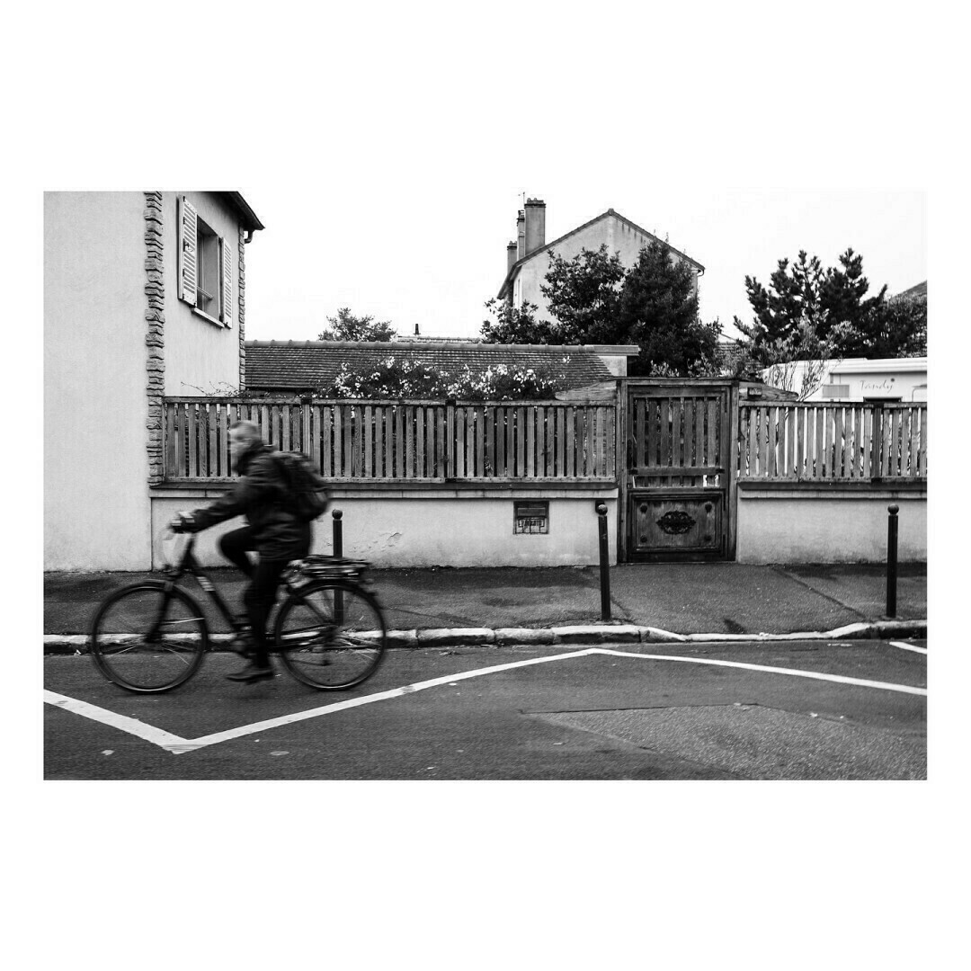 Une photo en noir et blanc montre un cycliste vêtu d’un manteau et d’un sac à dos roulant devant une maison. L’image est légèrement floue, capturant le mouvement du vélo. En arrière-plan, une clôture en bois et une maison aux volets fermés.