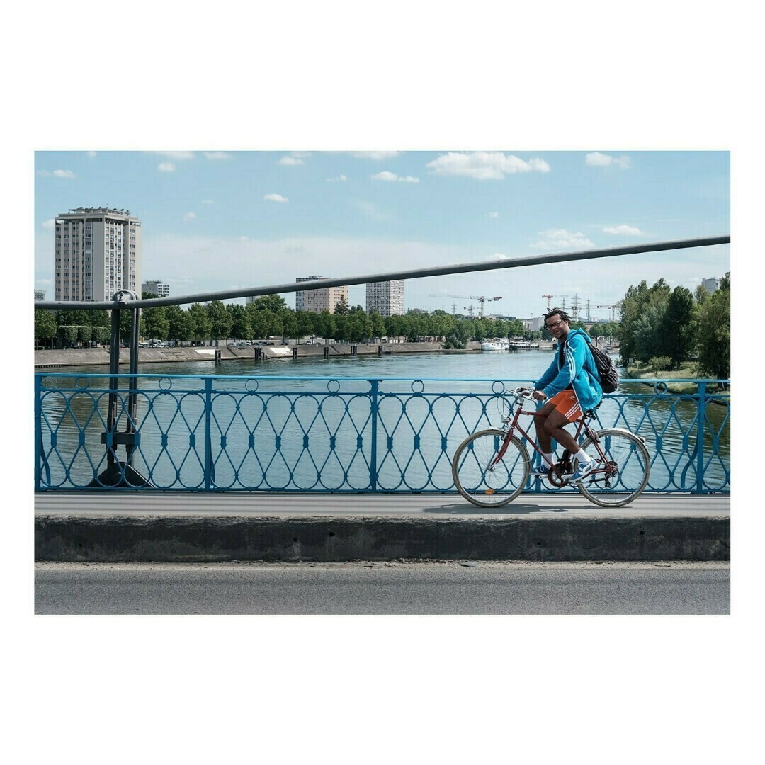 Un homme vêtu d’un short orange, d’un sweat bleu et d’un sac à dos noir traverse un pont au design métallique bleu, à vélo. En arrière-plan, une rivière, des immeubles et un ciel bleu parsemé de quelques nuages blancs.