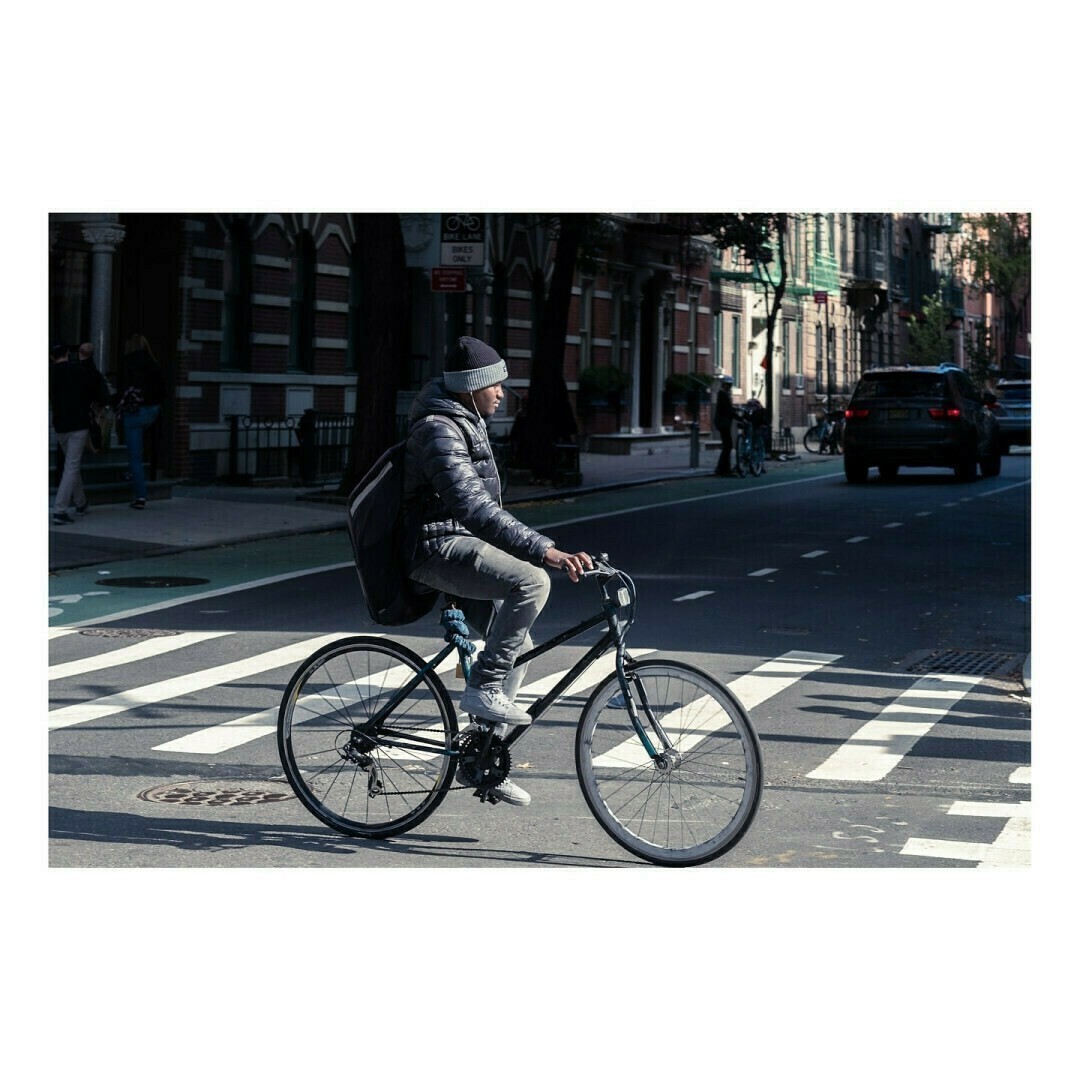 Un homme en doudoune, bonnet et jean roule sur un vélo de ville noir. Il porte un grand sac à dos et circule dans une rue avec des immeubles en briques et des voitures en arrière-plan.