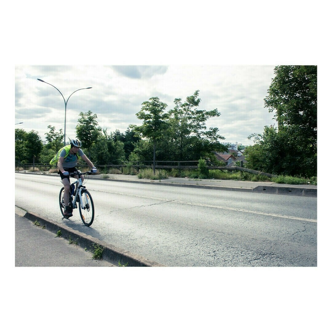 Un homme en tenue de cyclisme avec un casque et un sac à dos roule sur une route légèrement en pente. Le ciel nuageux et les arbres en arrière-plan ajoutent une ambiance de plein air.