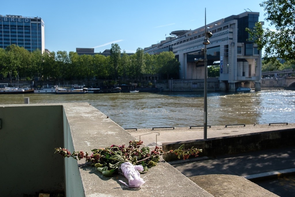 Une peluche et un bouquet fané sont posés sur un muret, face à la Seine ; en fond, l’imposant bâtiment du ministère de l’Économie.