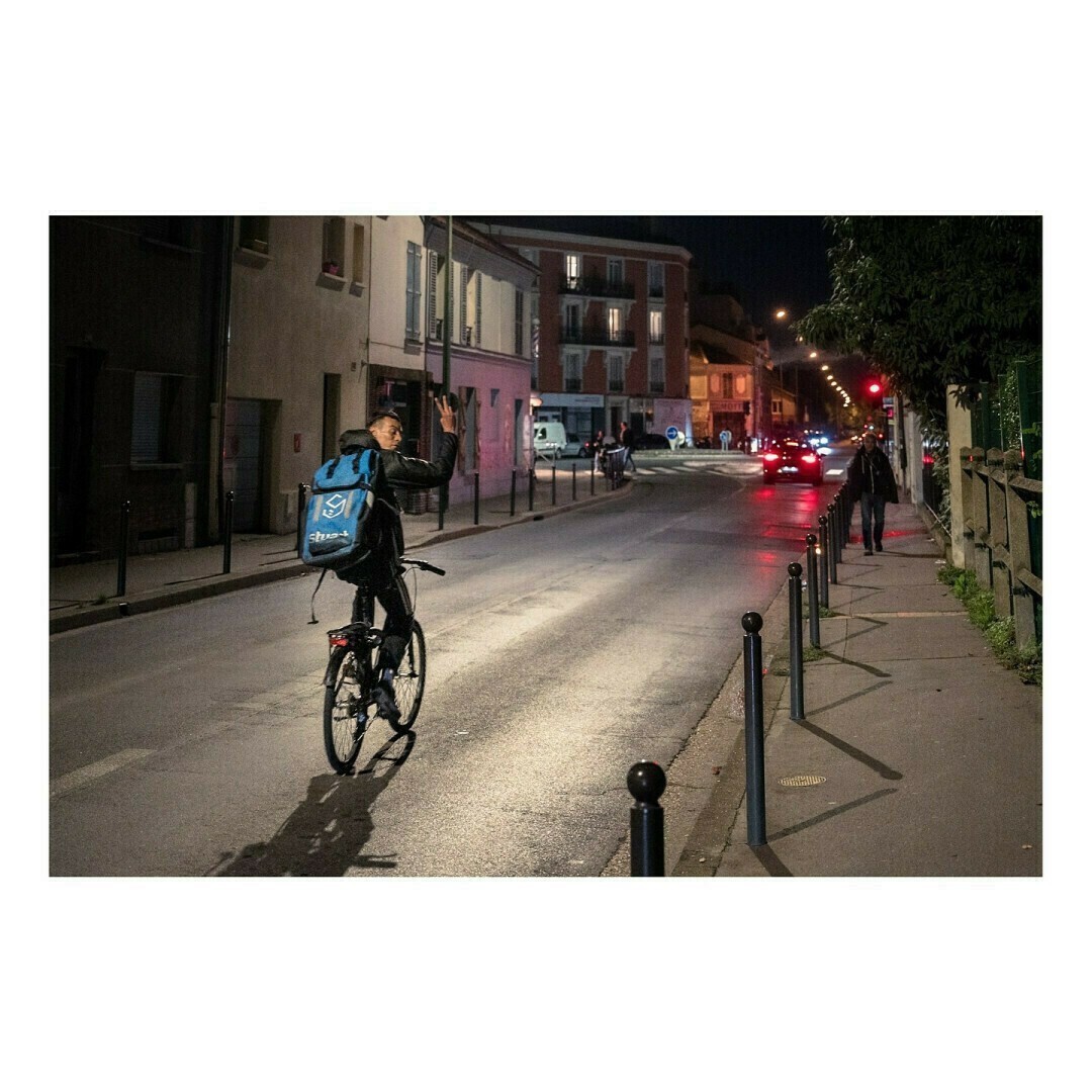 Un livreur en tenue sombre, avec un grand sac bleu de livraison sur le dos, roule sur une route éclairée par les phares des voitures et les lampadaires. Il lève la main comme pour me saluer.
