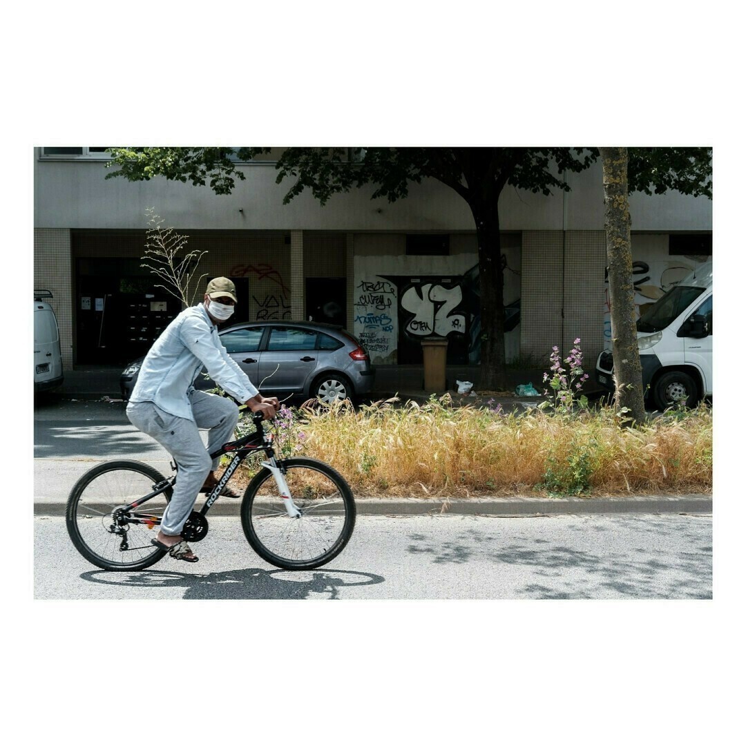 Un homme portant un survêtement gris, une casquette et un masque roule sur un VTT noir et blanc. Il circule sur une route bordée d’herbes hautes et de fleurs, avec en arrière-plan une façade d’immeuble taguée.