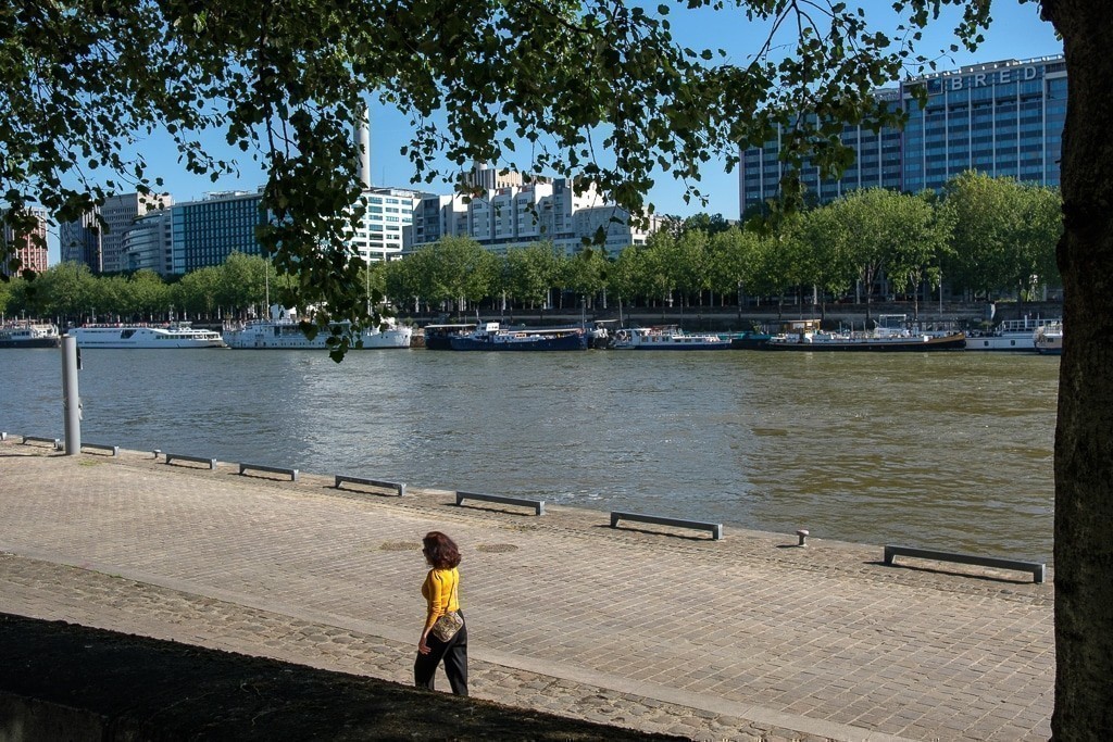 Une femme en haut jaune marche sur le quai ensoleillé, bordé d’arbres, avec la Seine et une rangée de bateaux à quai en arrière-plan.