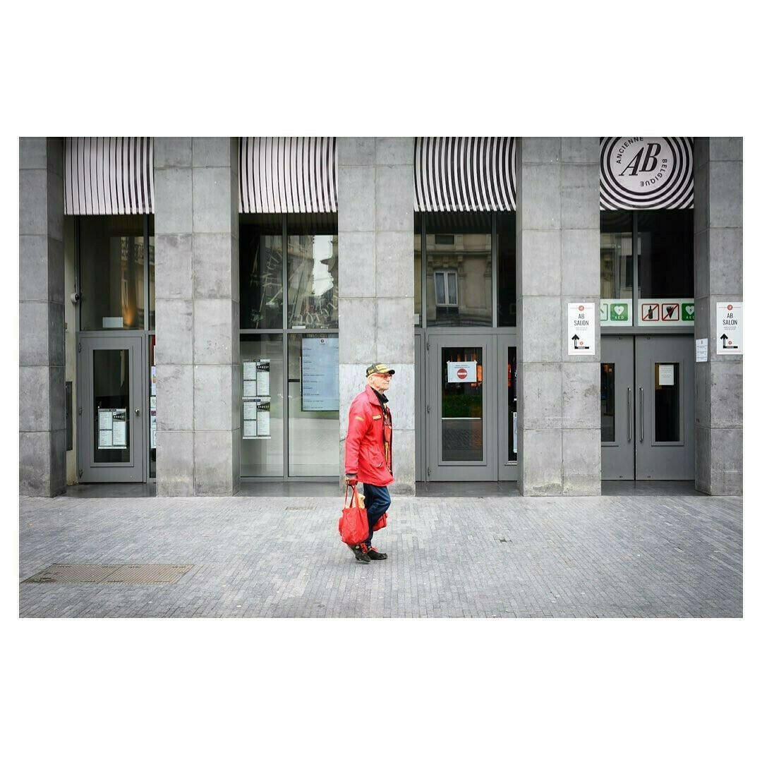 Un homme habillé de rouge passe devant une façade grise, graphique, contrastant ainsi avec les couleurs de l'arrière-plan, la scène se passe à Bruxelles en Belgique.