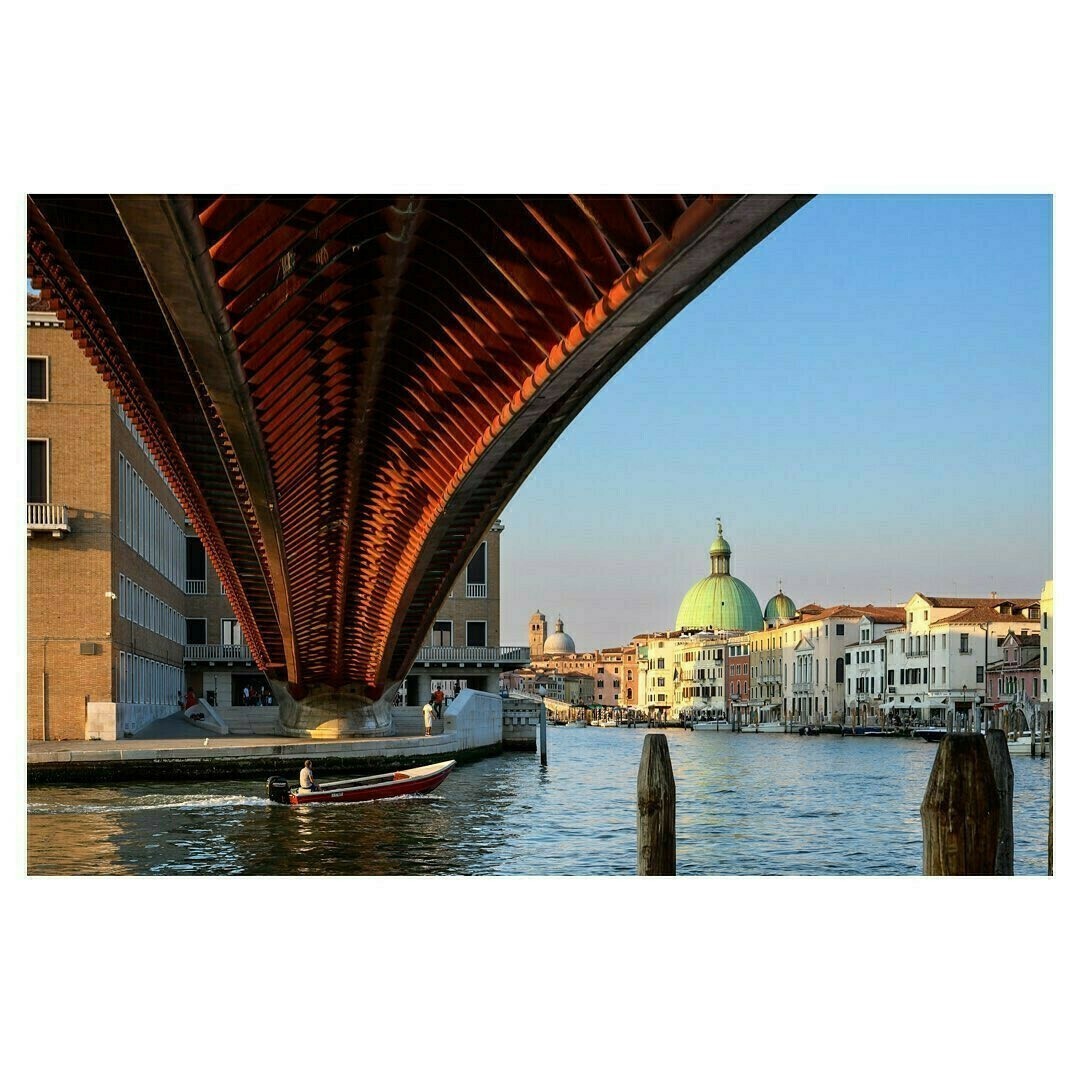 Cette photo montre une vue d'un canal à Venise, avec le pont de la Constitution (Ponte della Costituzione) au premier plan. La structure moderne du pont contraste avec l'architecture classique environnante, notamment le dôme vert de l'église San Simeone Piccolo visible au centre de l'image. Une barque navigue paisiblement sur l'eau au crépuscule, ajoutant une touche de vie à cette scène urbaine emblématique. Les teintes chaudes du pont éclairé par le soleil couchant apportent une belle harmonie à l'ensemble.