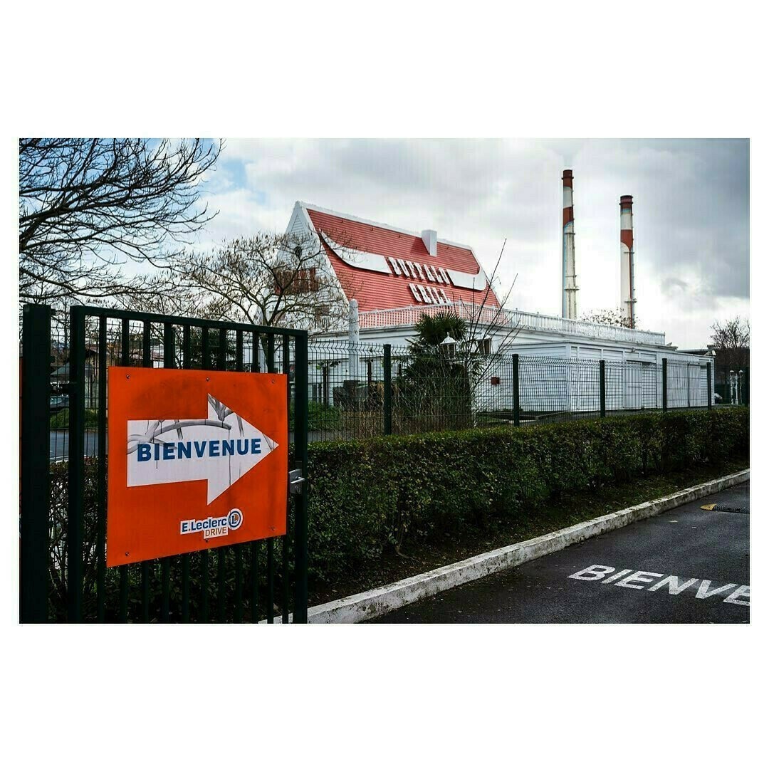 À Vitry-sur-Seine, cette scène capture le contraste frappant entre les éléments industriels et les marques de la vie de tous les jours. La pancarte accueillante d’un supermarché se juxtapose aux imposantes cheminées de l'ancienne centrale EDF voisine, reflétant une dualité urbaine où le fonctionnel rencontre le poétique. Un témoignage visuel de la cohabitation entre activité économique et vie locale.