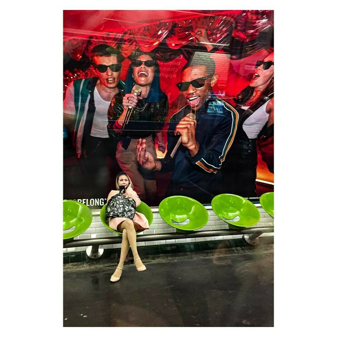 Cette photo capture une scène dans une station de métro. Au premier plan, une jeune femme est assise sur un banc en métal, constitué de sièges verts arrondis. Elle porte une robe légère, des collants couleur chair, et des chaussures élégantes. Elle est concentrée sur son téléphone portable, qu’elle tient d’une main, tandis que l’autre repose sur ses genoux.

En arrière-plan, un immense panneau publicitaire coloré occupe presque tout le mur. Il montre un groupe de personnes souriantes et énergiques, portant des lunettes de soleil et semblant chanter ou s’amuser avec des micros. Le contraste entre l’énergie exubérante du panneau publicitaire et l’attitude calme et isolée de la femme crée un effet visuel intriguant. 

L'éclairage de la station est typiquement artificiel, avec une légère brillance réfléchie sur les surfaces métalliques, accentuant les textures et les couleurs de la scène.