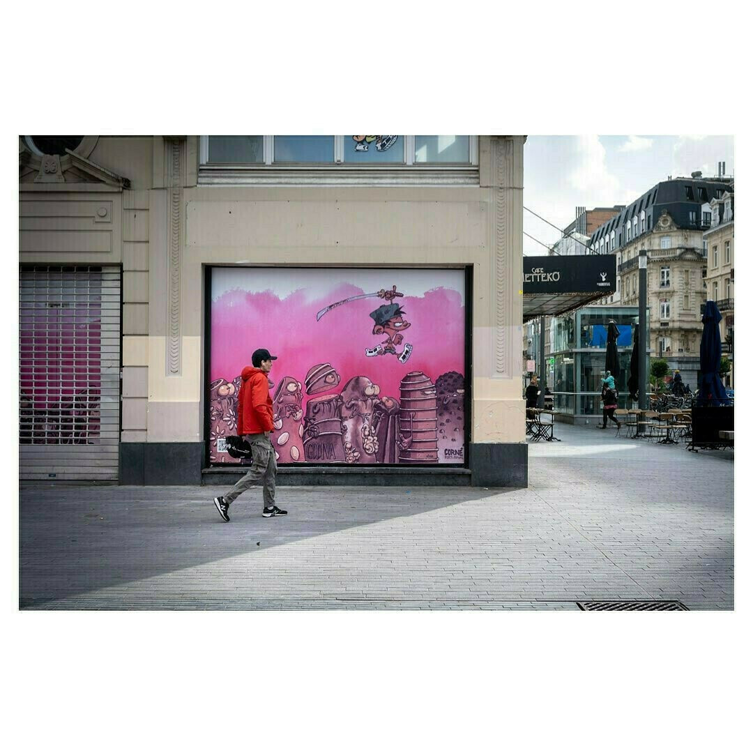 Dans une rue urbaine animée, un homme vêtu d’un sweat à capuche rouge et d’un pantalon kaki marche d’un pas décidé sur un trottoir pavé. Il porte une casquette noire et des baskets sombres, et tient un appareil photo à la main, suggérant qu’il est peut-être un photographe en action.

Derrière lui, une grande fresque murale attire l’attention. Elle représente un personnage de bande dessinée en train de sauter dans les airs, brandissant un sabre avec énergie. Le fond de la fresque est rose, contrastant vivement avec les tons plus neutres du bâtiment. En bas du mur peint, des robots et des créatures mécaniques sont alignés, ajoutant une dimension fantastique et urbaine à la scène.

L’environnement architectural est typiquement européen, avec des façades de pierre ornées et des grandes fenêtres. À droite, une terrasse de café avec des chaises et des tables est partiellement occupée, et quelques passants se déplacent en arrière-plan. Un abri en verre pour les transports en commun est visible, suggérant un centre-ville dynamique.

La composition de la photo joue avec les lignes architecturales et le mouvement du passant, créant un parallèle entre l’homme en rouge et le personnage bondissant de la fresque.