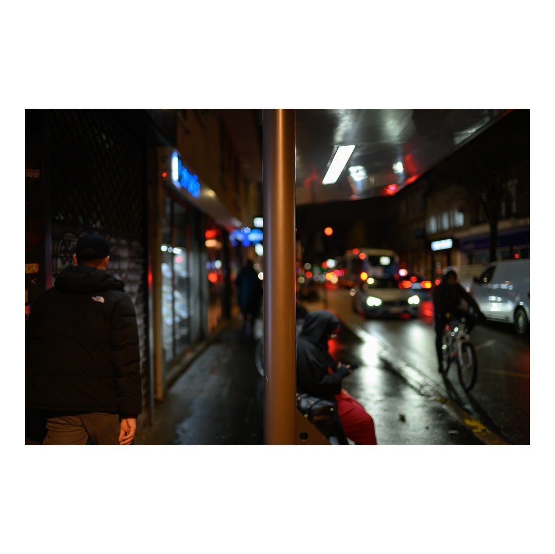 Rue animée de nuit avec un cadrage divisé par un poteau central. Sur la gauche, des passants marchent le long des vitrines éclairées, tandis qu'à droite, des cyclistes et des véhicules roulent sur une chaussée humide.