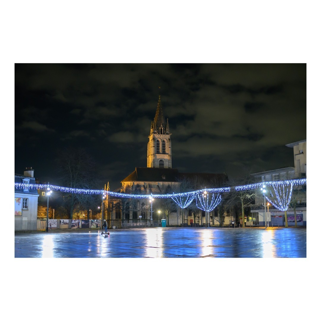 Une église se dresse majestueusement au centre de la place illuminée par des guirlandes lumineuses suspendues. Le sol reflète les lumières, créant une atmosphère paisible. Un enfant, sur une petite voiture, traverse l'espace vide.