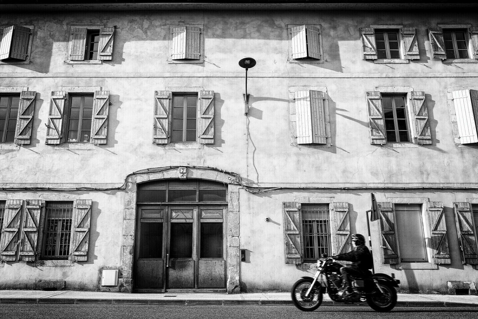 Un immeuble, vieux, blanc, avec une route devant, et une moto qui passe dessus.