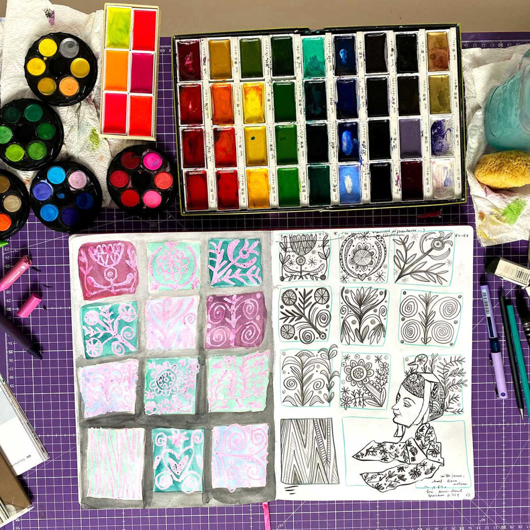 Carnet de croquis remplis de dessins inspirés par les motifs et costumes bretons. Du matériel artistique est visible : godets d’aquarelle, palette en céramique, pinceaux, crayons divers.