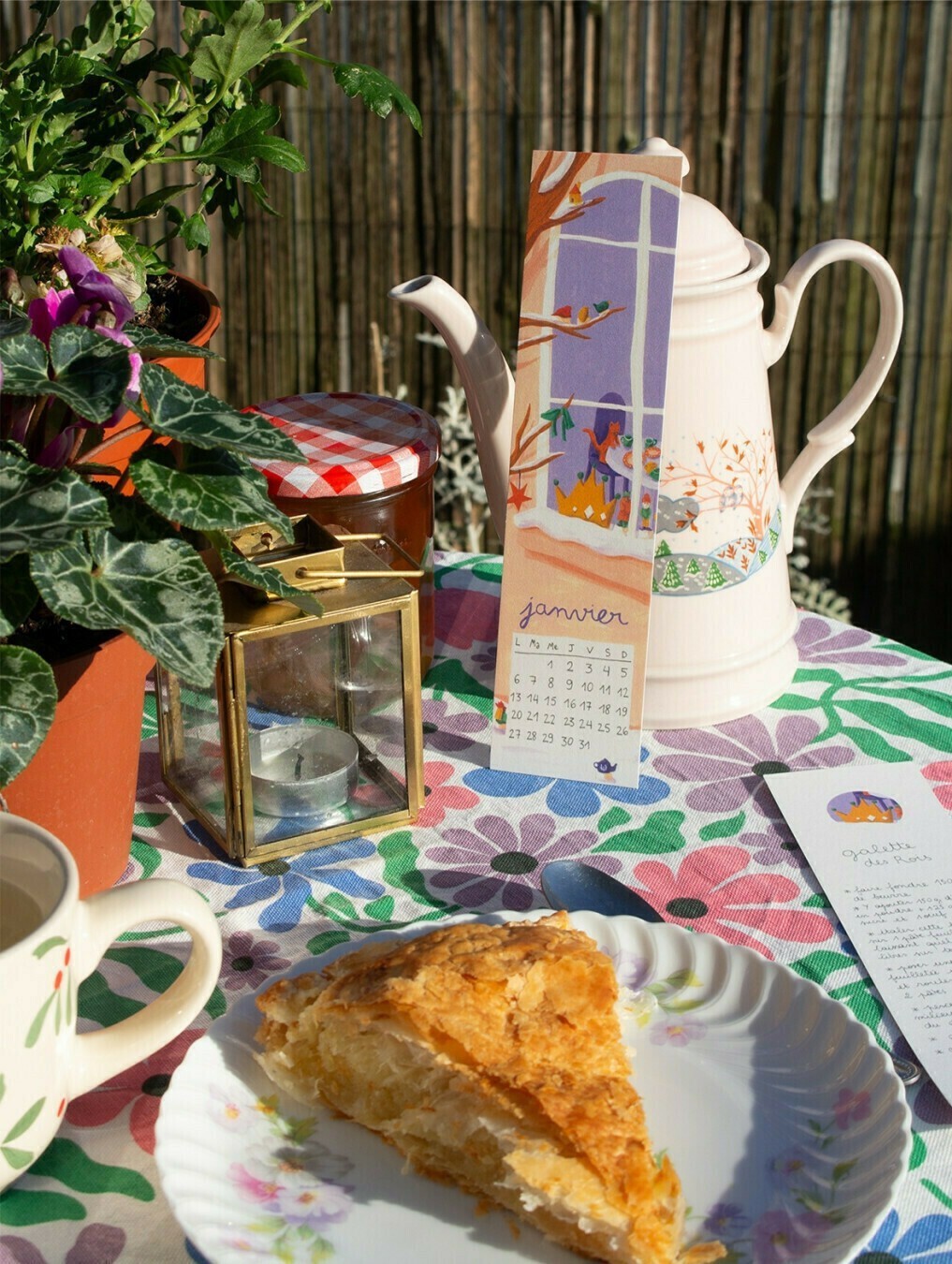 Photographie ensoleillée d'une table de jardin avec une nappe à fleurs, sur laquelle sont posées des plantes, une lanterne, une assiette avec une part de galette à la frangipane, une tasse de thé, et la page de janvier du calendrier 2025 illustré par Mathilde Villerot