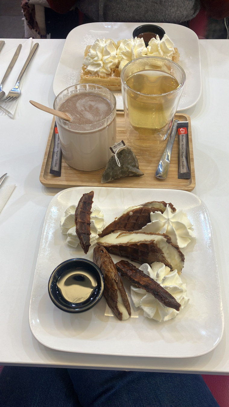 Assiette carrée avec des lacquemant à coupés en petites tranches. Glace à l’intérieur, chantilly dans chaque coin. Une coupelle avec le coulis. Au second plan un plateau avec un thé et un chocolat chaud. Arrière plan, assiette carrée avec gaufre de Bruxelles, banane et chantilly au dessus
