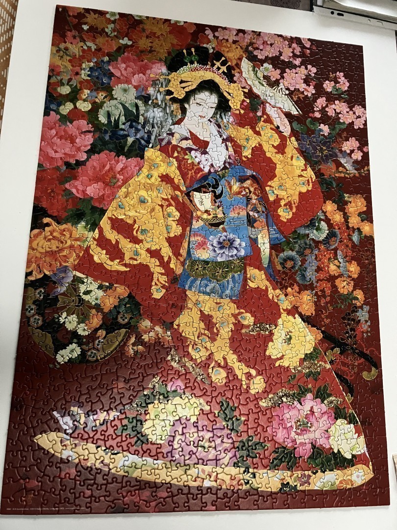 Puzzle finalisé. Jeune femme japonaise habillée d’un kimono fleuri. Fond rouge avec fleurs roses et mauves. Obi bleu. Elle est entourée de fleurs autour de sa tête