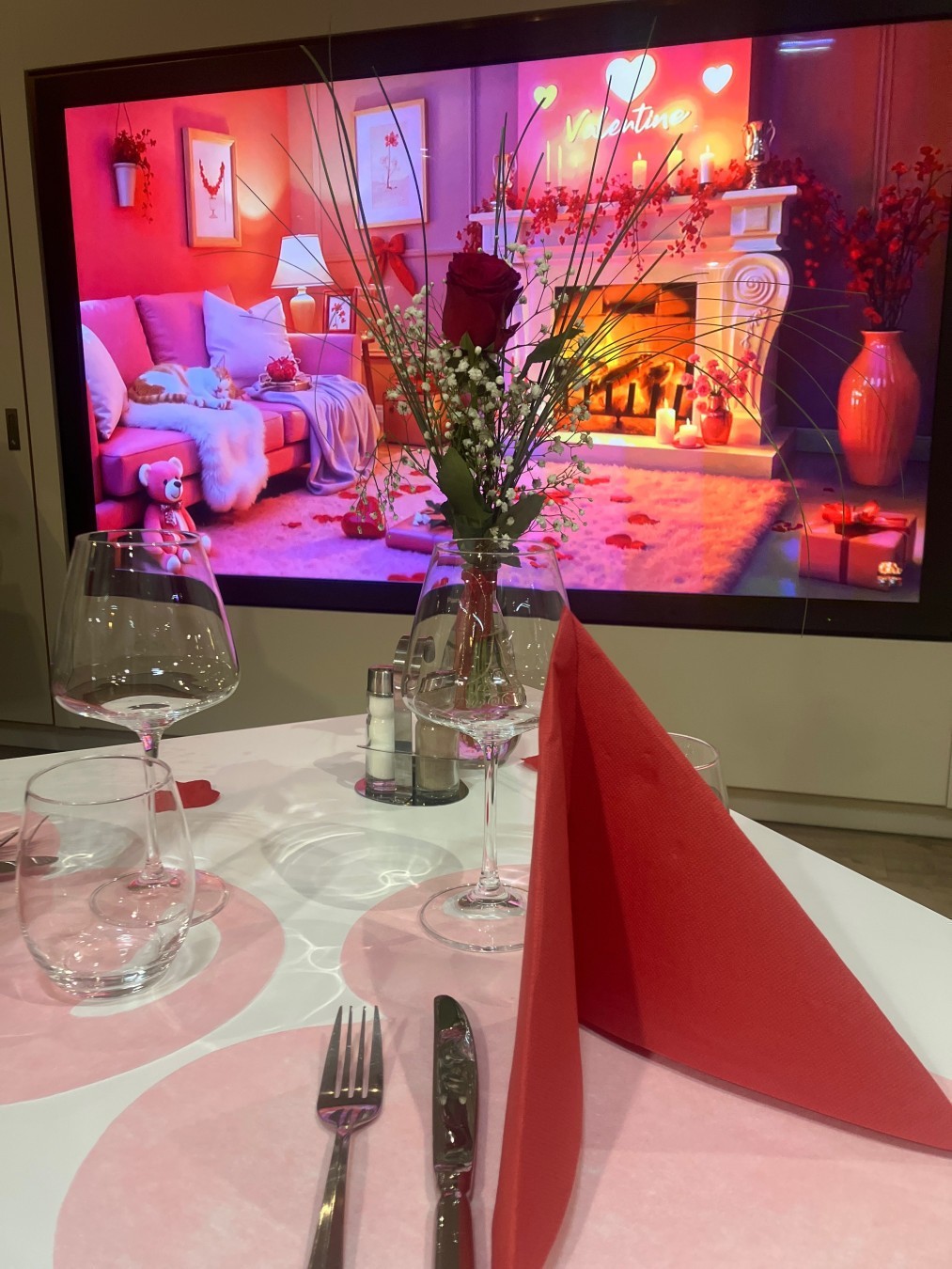 Une table blanche décorée avec un set de table papier rose en cœur et une serviette rouge. Le fond est une image d’un salon avec feu de cheminée. Ton rose