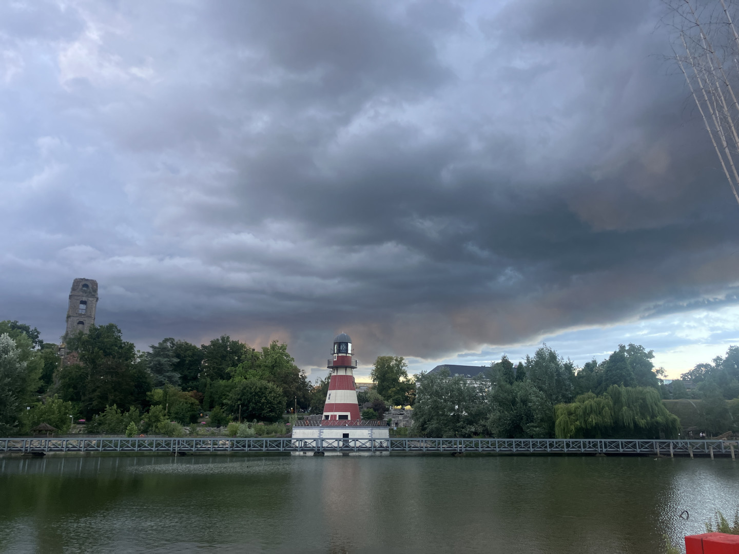 un paysage avec un plan d'eau au premier plan. Au centre de l'image, il y a un phare de couleur rouge et blanche situé sur une petite île ou une jetée. À gauche du phare, on peut voir une tour en pierre partiellement cachée par des arbres. Le ciel est couvert de nuages sombres et menaçants, suggérant un temps orageux ou une tempête imminente. Les arbres et la végétation autour du plan d'eau sont verts et denses, ajoutant une touche de nature à la scène. Une passerelle en métal traverse l'eau, reliant les deux rives et passant près du phare.