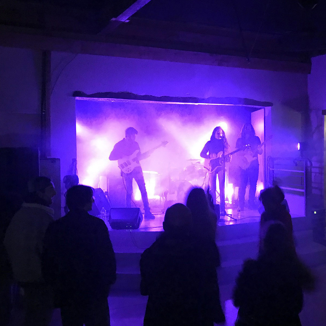 le goupe de rock Frank joue sur une petite scène. Le public est de dos, la lumière est violette. la chanteuse devant joue de la guitare électrique et chante, sur sa droite et sur sa gauche deux guitaristes jouent. Au fond on distingue une batterie, mais on ne voit pas le batteur, il y a de la fumée.