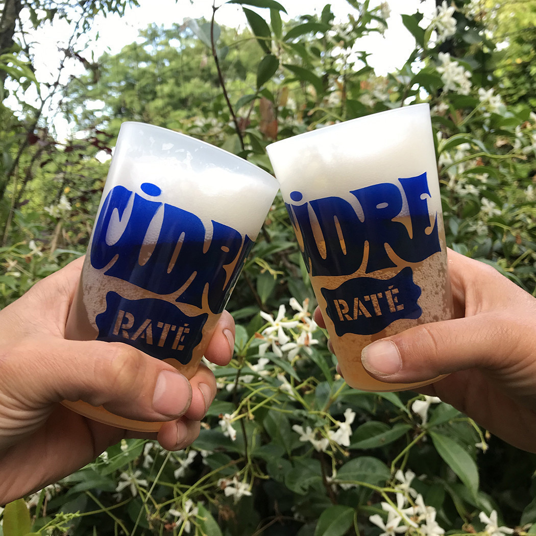 Deux mains qui trinquent avec des ecocups transparents remplis de bière aux deux tiers. Sur les écocups, est écrit en bleu foncé : cidre raté.
Les lettrages ont étés dessinés par Bon Pour 1 Tour en 2024. En fond de l'image des arbres et les branches fleuries d'un faux jasmin.