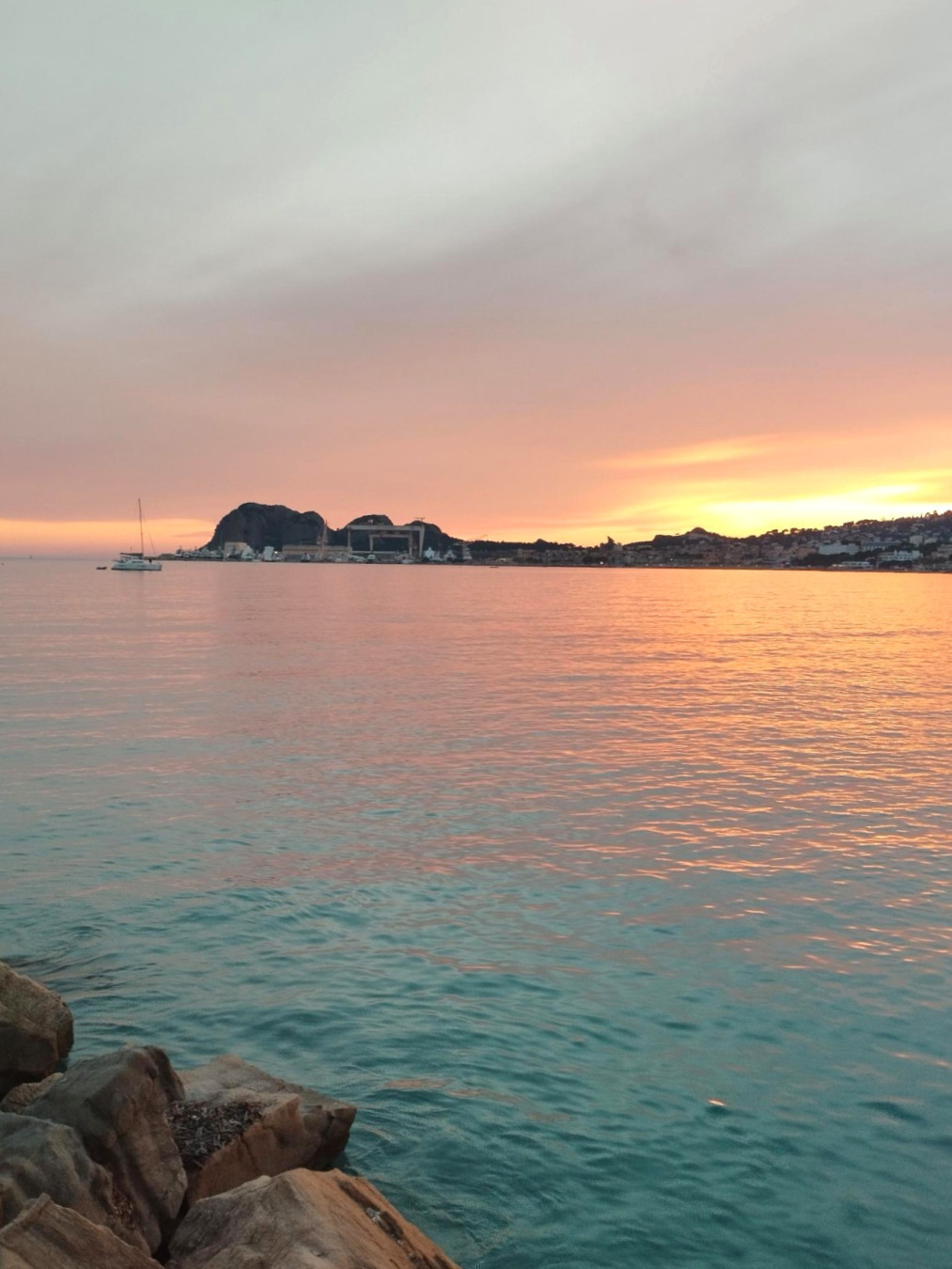 La Ciotat, vu sur la mer, au loin les  falaises de la calanque de Figuerolkes. Le soir tombe, lumière diffuse de rose, orange, vert d'eau, jaune, des couleurs pastels. on voit surtout la mer et le ciel. La mer est calme et il n'y a pas de nuage, pas de bateau. On voit quelques rochers en bas à gauche de la digue de laquelle la photo a été prise. Cela donne un sentiment de douceur, bien être, sérénité,