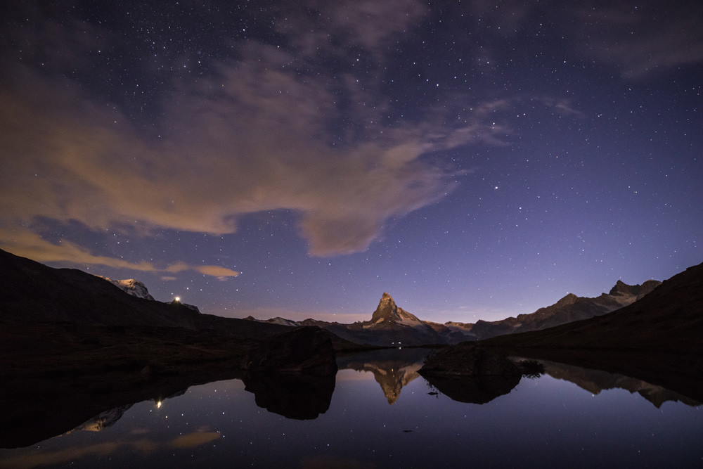 Photo d'un paysage de montagne nocturne.
Le Cervin se reflète dans un lac sous les étoiles. La partie haute de la montagne est quelques autres sommets sembles éclairés, sans doute par la lune, le reste du paysage est dans le noir à l'exception des eaux du lac qui reflètent le ciel étoilé. 
Il y a quelques nuages dans le ciel, mais qui n'altèrent pas l'impression de beau temps.
Sur une montagne à gauche on voit une lumière qui brille fortement, sans doute une gare de téléphérique. On distingue aussi des points lumineux sur les flancs du Cervin, probablement des refuges.
-
Photo of a mountain landscape at night.
The Matterhorn is reflected in a lake under the stars. The upper part of the mountain and a few other peaks seem to be illuminated, probably by the moon, while the rest of the landscape is in darkness, except for the waters of the lake, which reflect the starry sky. 
There are a few clouds in the sky, but they don't alter the impression of fine weather.
On a mountain on the left, we see a light shining brightly, probably a cable car station. We can also make out bright spots on the flanks of the Matterhorn, probably huts.