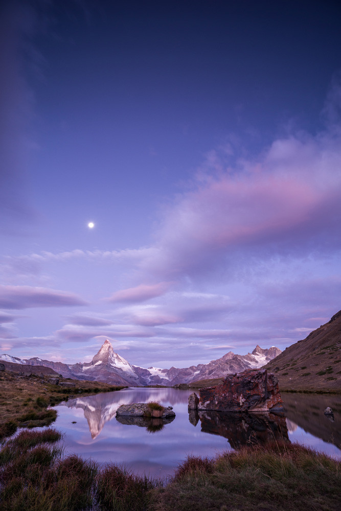 Photo d'un paysage de montagne au lever du jour, l'image est au format portrait.
Le Cervin se reflète dans un petit lac de montagne d'où émergent deux rochers. Les berges du lac sont herbeuses.
Le ciel occupe les deux tiers supérieurs de l'image, la moitié basse est envahie de nuages rose pastel, la partie haute vire au bleu sombre. Au milieu, côté gauche, la pleine lune domine le Cervin. 
L'ambiance est très douce.
-
Photo of a mountain landscape at sunrise, the image is in portrait format.
The Matterhorn is reflected in a small mountain lake from which two rocks emerge. The banks of the lake are grassy.
The sky occupies the upper two-thirds of the picture, the lower half invaded by pastel-pink clouds, the upper part turning dark blue. In the middle, on the left, the full moon dominates the Matterhorn. 
The atmosphere is very soft.